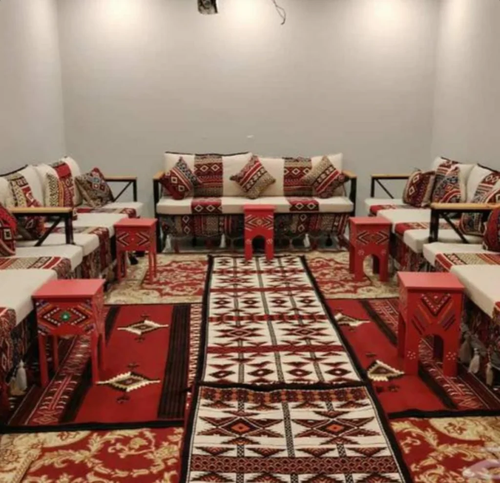كنب مجلس بنقوش تراثية حمراء مع وسائد وطاولات ضيافة فوق سجاد منقوش.