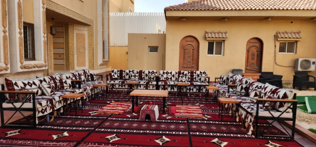كنب تراثي يحيط بسجاد أحمر وطاولات ضيافة في فناء بجوار مبنى.