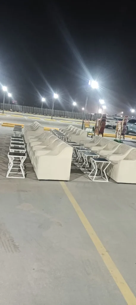 كنب أبيض وطاولات موزعة في صفوف داخل مساحة مناسبة خارجية مضاءة ليلاً.
