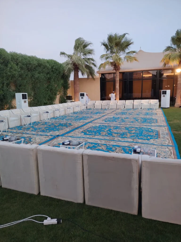كنب فاتح اللون يحيط بسجاد أزرق في مساحة خارجية بين الأشجار والنخيل.
