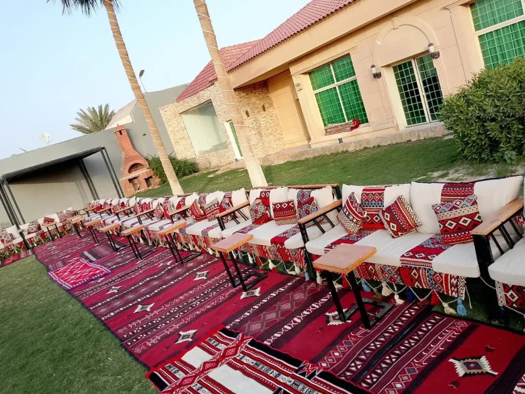 كنب تراثي موزع على سجاد أحمر في حديقة خارجية مع طاولات ضيافة.